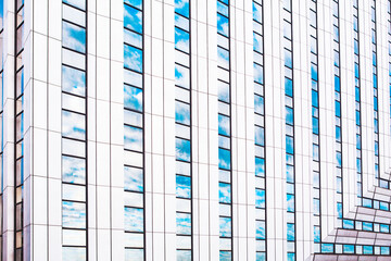 Modern high rise building facade features repeating vertical white panels and reflective glass windows. Blue skies and clouds are mirrored perfectly in glass, creating minimalist, abstract pattern