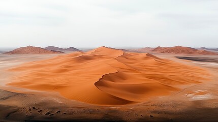 Fototapeta premium Vast expanse of rolling sand dunes in vibrant orange and brown hues, with distant mountains under a hazy, overcast sky. The light is soft and diffused.