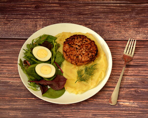 A fried meat patty with mashed potatoes, a sprig of dill, and lettuce leaves with a sliced ​​boiled egg on a wooden table, with a fork nearby.