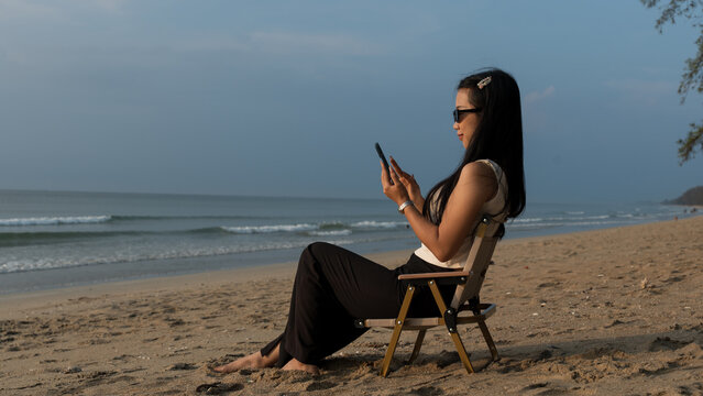 Woman using a smartphone while relaxing on a beach chair beside the quiet ocean. - Powered by Adobe