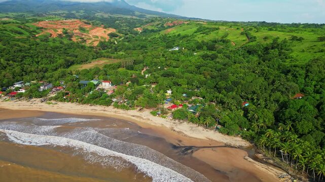 Closing‑in aerial approaching the coastline town, revealing sandy shoreline, gentle waves, and colorful rooftops framed by lush tropical scenery at Quinawan Beach in Mariveles, Bataan, Philippines.