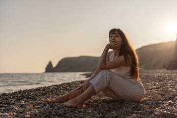 Woman, beach, sunset. Serene woman sits thoughtfully on a tranquil pebbled beach at golden hour, ocean waves gently lapping, with ample copy space.