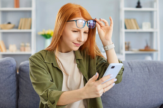 Young girl wearing eyeglasses in casual clothes using mobile phone sitting on sofa at home. Woman with vision problems holding cellphone reading sms message. Farsightedness and presbyopia concept.