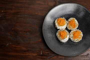 Close-up of four steamed dimsum topped with grated carrot, arranged on a dark plate over rustic wooden background. Ideal for Asian food and menu concepts.