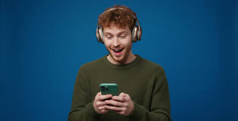 Young man with headphones smiles while looking at a smartphone, capturing a relaxed and cheerful digital moment that reflects enjoyment of music, social media, or entertainment indoors.
