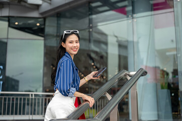 Fototapeta premium Asian woman holding phone and carrying shopping bags while walking upstairs to a mall's glass door