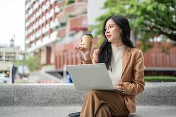Pretty asian woman office worker holding coffee cup over laptop sitting crossed leg on bench in park