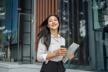 Happy asian woman office worker holding coffee cup carrying laptop and walking outside mall building