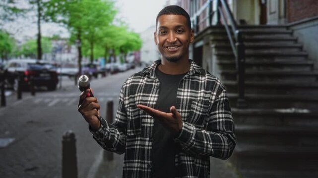 Young man holding electric screwdriver in right hand and presenting it with left palm up on a street in front of building steps; confident diy repair mood.