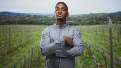 Man with arms crossed wearing a buttoned shirt standing in a vineyard forest with trellis rows and distant hills; confidence resilience.