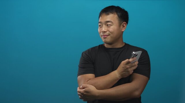 Young chinese man smiling while holding smartphone with both bare hands at chest level in turquoise studio setting; serenity.