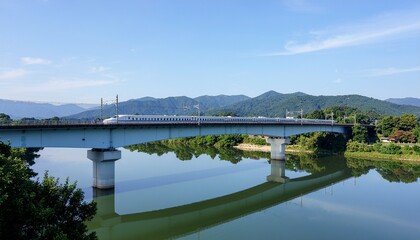 Fototapeta premium A train travels across a bridge over a river, with mountains visible in the background under a clear sky.