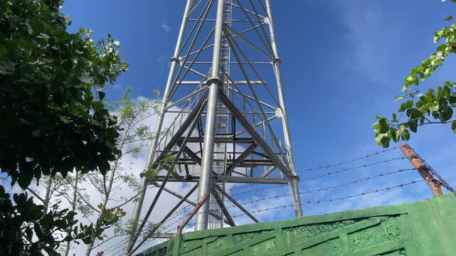 Metal communication tower rises above tropical leaves on Mount Magarwak with steel lattice structure framed against a clear blue sky