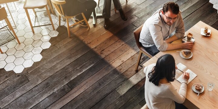 Aerial view of a cafe with wooden floors. Two people sit at a table with coffee and pastries. Cozy cafe atmosphere with wooden decor and relaxed vibe.