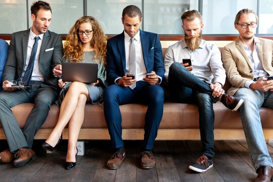 Group of professionals sitting on a couch, using laptops and phones. Business attire, diverse group, engaged in technology and communication. Marketing professionals using devices, sitting in a row.