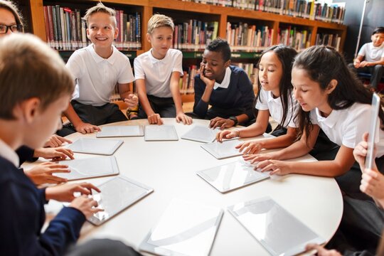 Group of diverse students in a library using tablets. Kids in school uniforms engaged in learning. Education and technology in a classroom setting. Diverse elementary school kids in library.