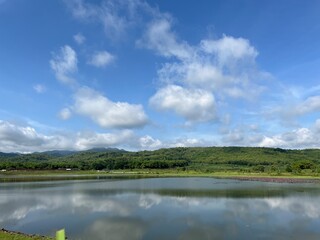 Bright reservoir landscape with blue sky, scattered clouds, and rolling greenery under tropical sunlight.