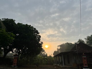 Early morning sun rising behind trees and rural houses in a quiet tropical village scene.