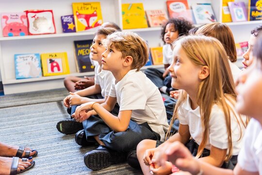 Children sitting on the floor in a classroom, listening attentively. Books on shelves in the background. Kids engaged in a learning environment. Elementary school children sitting on the floor.