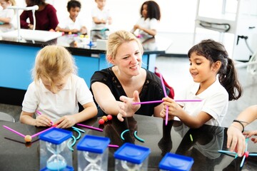 Female teacher with blonde hair helping diverse children with STEM activities. Children engaged in STEM, building with sticks. STEM learning in a classroom setting. Elementary school education.