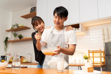 Young couple cooking together in kitchen, man whisking batter in bowl while woman embraces him from behind with happy expression, modern home interior with wooden shelves and white cabinets