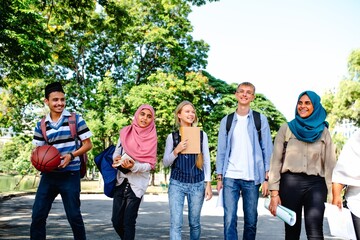 Diverse group of young adults walking outdoors, carrying books and a basketball. Smiling, wearing casual clothes, enjoying a sunny day together. Diverse students walking together after school.