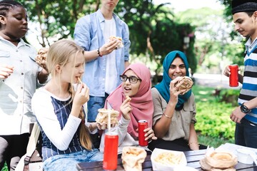 Group of diverse friends enjoying a picnic outdoors, eating sandwiches and drinking soda. Multicultural gathering with food, laughter, and nature. Diverse friends having picnic in park.