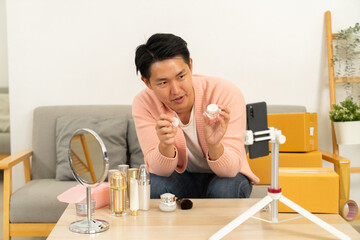 Man demonstrating skincare cream on camera for online product review, sitting on sofa with cosmetic bottles, mirror, and packaging boxes in cozy room