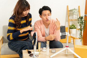 Young couple recording skincare product review with smartphone, applying facial mask patch and discussing beauty routine at home, cozy living room setting with makeup items on table