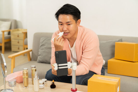 Young man applying lip balm while recording video with smartphone on tripod, surrounded by cosmetic products and cardboard boxes, cozy home setting