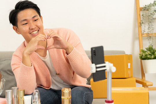 Young man recording video with smartphone for online selling product review, smiling and making heart shape with hands, surrounded by cosmetic bottles and cardboard boxes