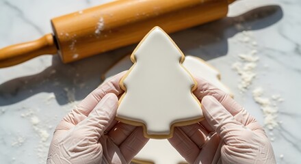 Baker's hands in gloves holding a freshly decorated Christmas tree cookie with white icing, with a rolling pin in the background.