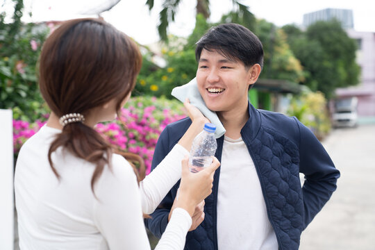 Young man smiling while woman holds water bottle and cools his face with ice pack outdoors in garden with flowers and trees