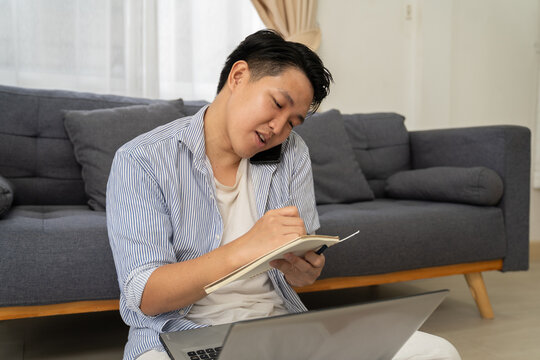 Man writing in notebook talking on phone working from home with laptop on floor in living room