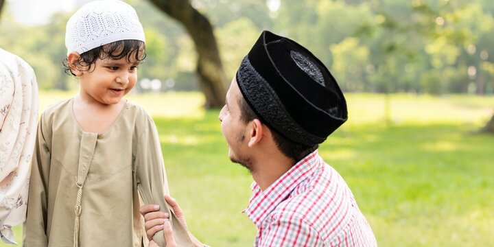 A muslim man and a young boy in traditional attire enjoy a sunny day in the park. The boy wears a white cap, and the man wears a black cap, both smiling warmly. Happy muslim family outdoors in a park. - Powered by Adobe