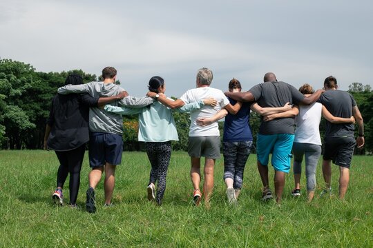 Diverse people hugging each other. Diverse group of men and women embrace each other at park. Unity and teamwork. Backside of friends standing in row huddle on green grass. Group of friends huddle