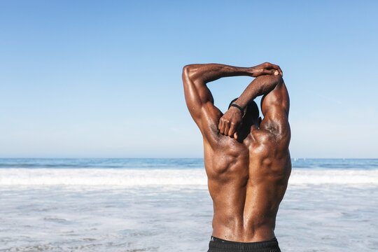 Man stretching on beach, ocean view. Muscular man, back view, stretching arms. Beach, ocean, stretching, exercise, fitness. Sunny day, ocean, beach. African American man doing exercise workout.