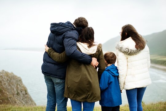 Family stand and huddle together in scenic landscape. Mother, father and children enjoys the outdoors together. Back view of family look coastal landscape. Family hugging each other looking at view