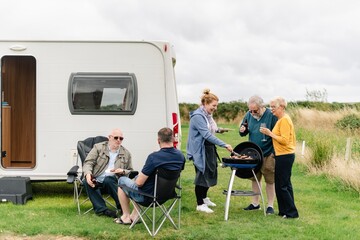 Diverse group of senior friends enjoying barbecue by a caravan. Outdoor gathering with friends. Relaxing near a caravan, cooking and chatting. Casual outdoor senior friends barbecue scene.