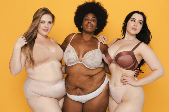 Three women in lingerie pose confidently against a yellow background, showcasing body positivity and diversity in body types and skin tones. Diverse women in lingerie, body positivity and inclusivity.