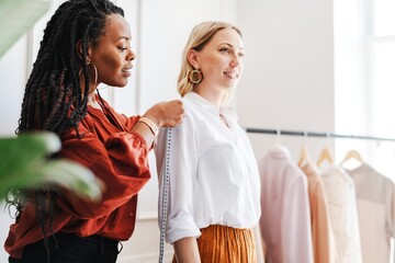 A Black stylist woman fitting outfit to Caucasian woman model in a stylish clothing store. The Black woman is helping the White woman with fitting, showcasing outfit fashion and personal styling.