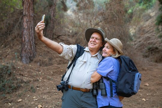 Elderly couple hiking, taking a selfie in the woods. Both wear hats and backpacks. Nature, adventure, and togetherness in a forest setting. Eldely couple taking selfie in forest while camping. - Powered by Adobe