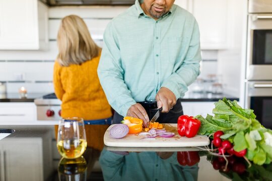 Man preparing meal and chopping vegetables in kitchen. Bell pepper, onion, and greens. Woman in background. Man cooking healthy menu with vegetables in kitchen with woman. People cooking in kitchen - Powered by Adobe