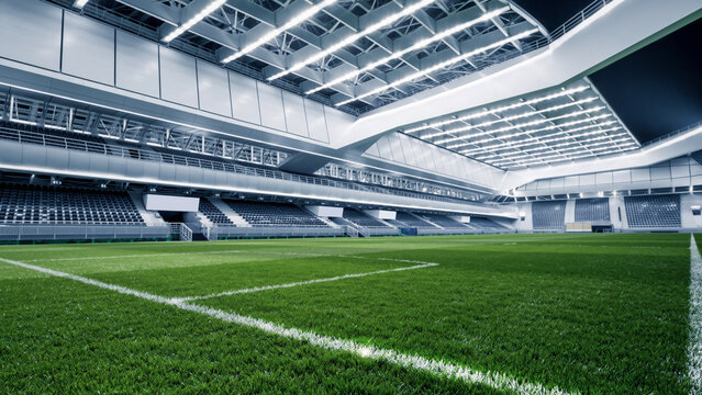 Empty modern football stadium with green grass field and rows of spectator seats under a bright roof.