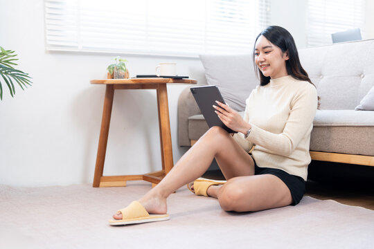Asian woman sitting on the floor in her living room, comfortably using a digital tablet for reading or streaming content at home.