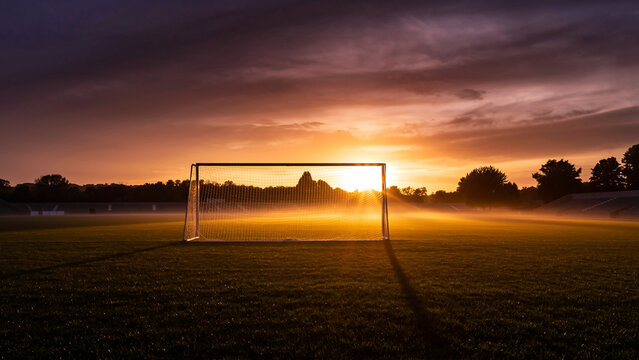 A soccer goal stands on a misty field at sunrise, casting a long shadow under a dramatic sky. - Powered by Adobe