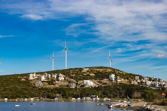 Modern Coastal Residences Sprawling Across a Green Hillside Topped with Tall Wind Turbines, Overlooking a Calm Bay with Small Boats Under a Clear Blue Sky. - Powered by Adobe