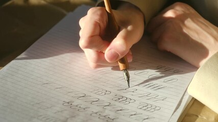 Close up view of a hand meticulously practicing lettering and penmanship exercises on lined paper with a vintage dip pen and ink, illuminated by warm sunlight from a nearby window