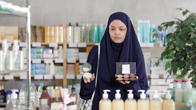 Muslim woman in traditional dress looking with interest at compact powder and foundation shades, carefully matching products for natural daily makeup at local drugstore