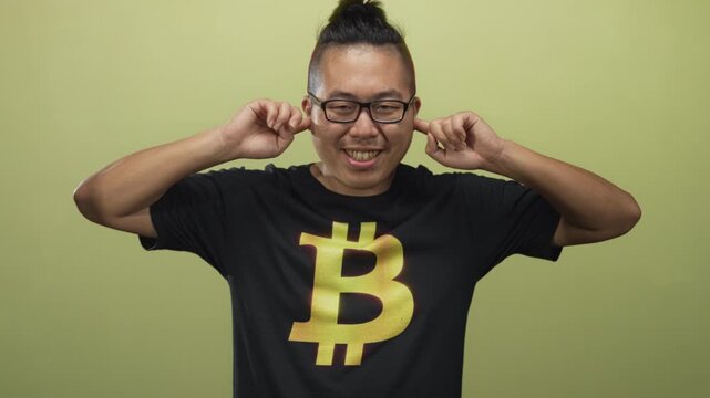 Man wearing bitcoin t shirt plugs fingers in ears in studio with olive green backdrop; annoyance silence.